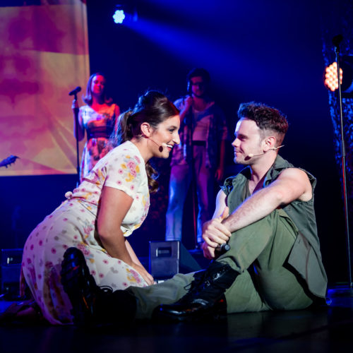 Female performer kneeling beside a seated soldier during an intimate stage scene