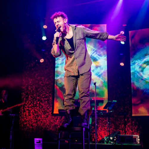 Male vocalist singing from a chair beneath vivid purple and red lighting