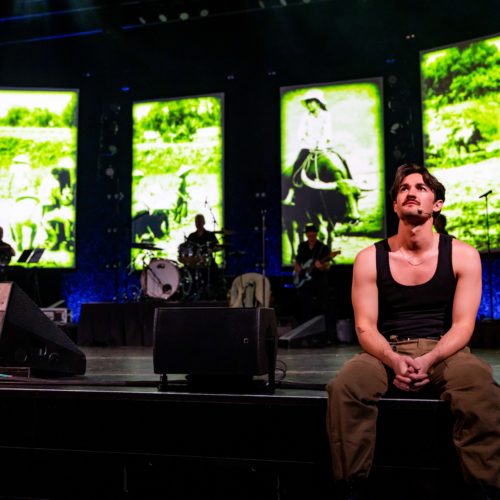 Male performer seated at the front of the stage before Vietnam War imagery