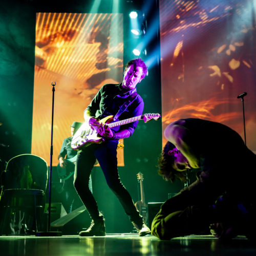 Electric guitarist performing beside a crouched cast member under green stage light