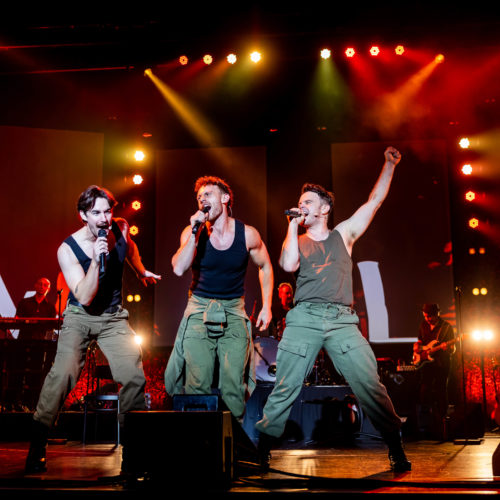 Three male vocalists singing together at the front of the stage