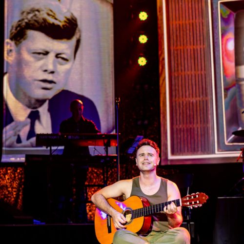 Seated male performer playing acoustic guitar before projected footage of John F. Kennedy