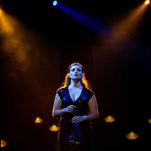 Female vocalist singing alone beneath blue and amber spotlights