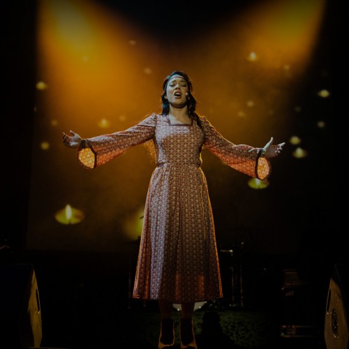 Female vocalist singing with her arms outstretched beneath amber spotlights